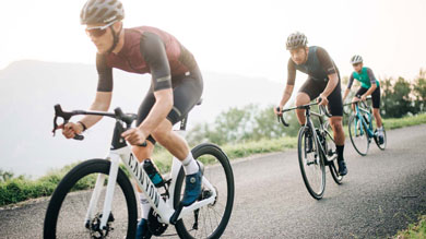Three cyclists wearing helmets ride on a paved road in a scenic outdoor area.