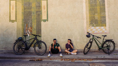 Two people sitting on a sidewalk, eating and drinking with bicycles parked nearby against a wall with murals.