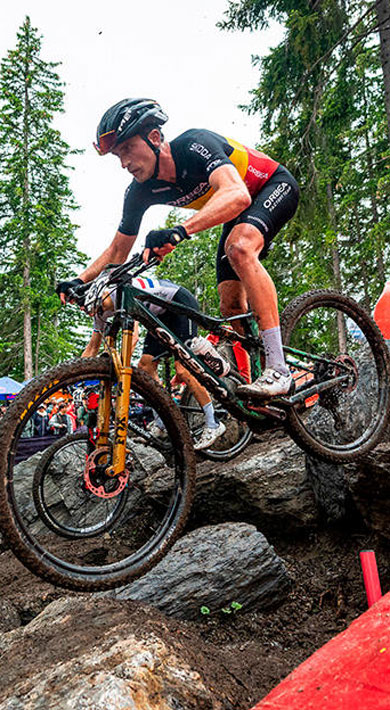 Cyclists navigating a rocky trail during a mountain bike race in a forest with spectators watching.