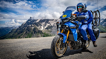 Motorcyclist in blue gear rides on a mountain road with stunning alpine scenery in the background.