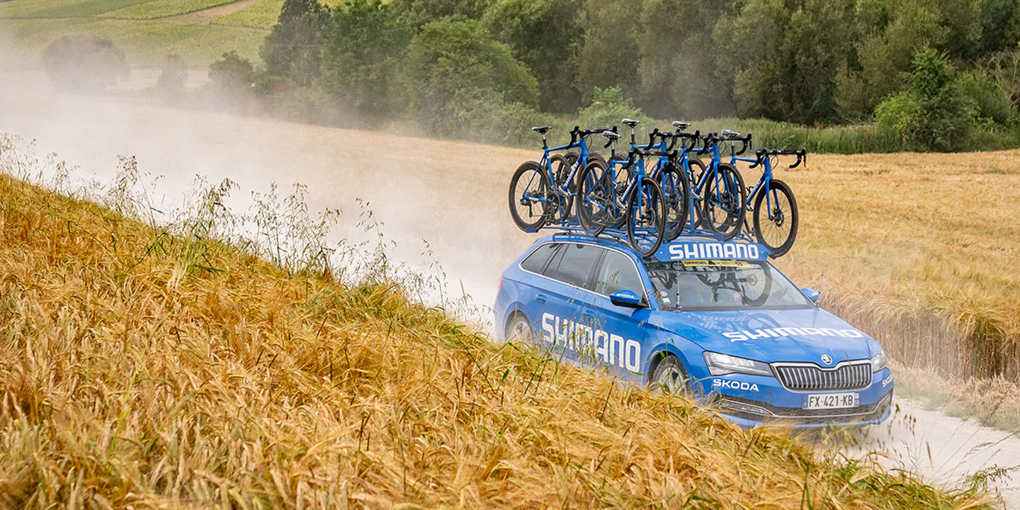 Blue Skoda support car with bicycles on roof driving on dirt road through fields
