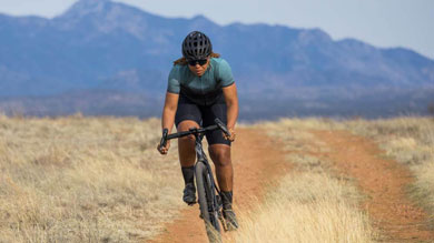 Cyclist riding on a dirt path through a grassy field with mountains in the background