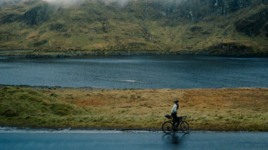 Cyclist rides along a calm lake with fog-covered mountains in the background