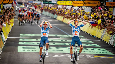 Deux coureurs célèbrent leur victoire en franchissant la ligne d'arrivée d'une course, avec des spectateurs en liesse en arrière-plan