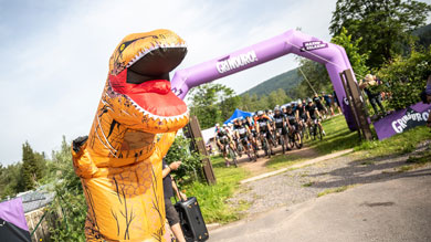 Person in dinosaur costume at the starting line of a bike race under a purple arch.